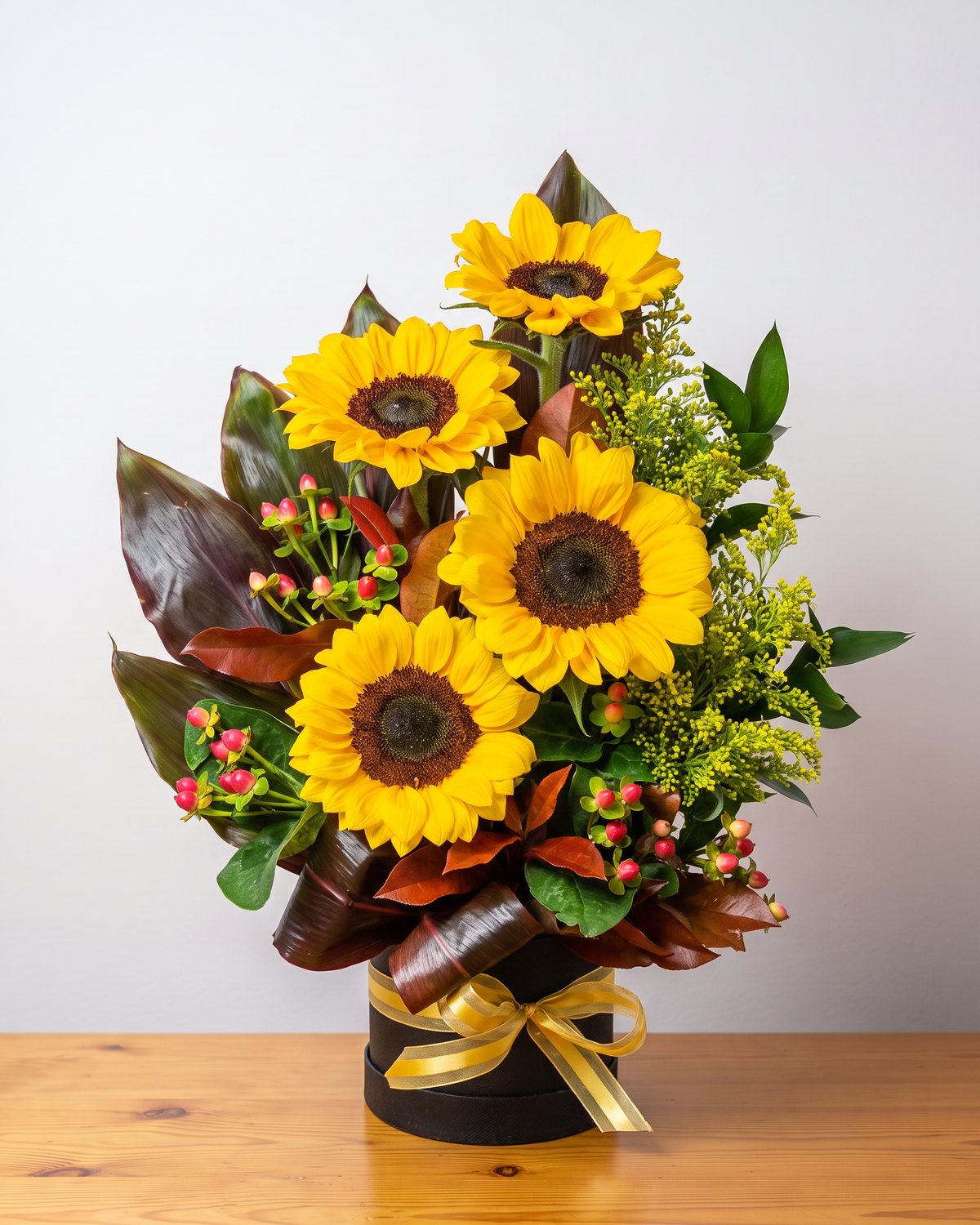 O box de girassol da casa: quatro girassóis subindo em diagonal entre folhas cor de vinho, solidago e hypericum, numa caixa redonda preta com laço de fita dourada listrada.