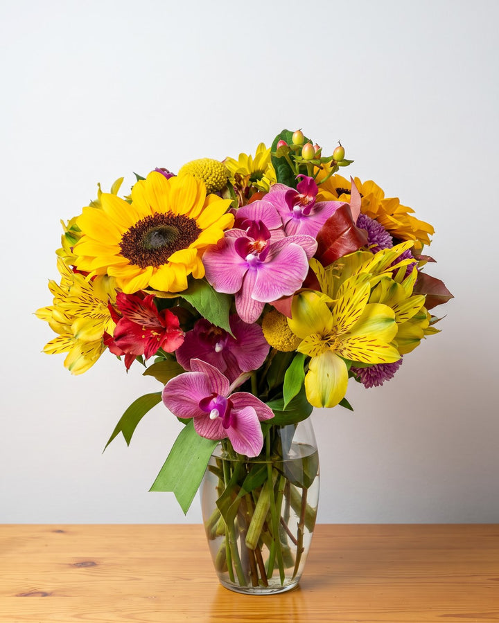 Arranjo mix de flores: girassol, orquídeas magenta, alstroemérias, craspédia e hypericum num vaso baixo de vidro