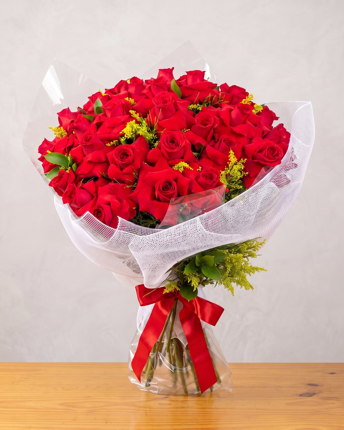O buquê de 36 rosas colombianas da casa: rosas vermelhas de pétalas pontudas com solidago, em celofane e tela branca enrolada, laço de cetim vermelho e saco de água, segurado por uma mão.