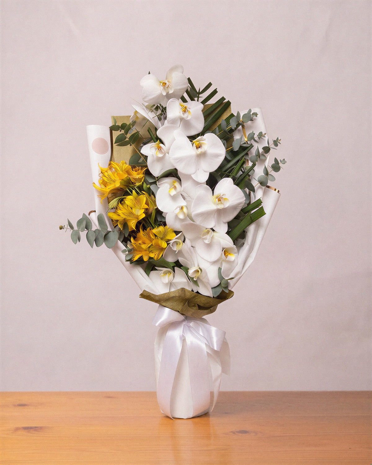 Buquê de orquídeas brancas em cascata com alstroemérias amarelas, folhas de palmeira e eucalipto, em papel branco com laço de cetim