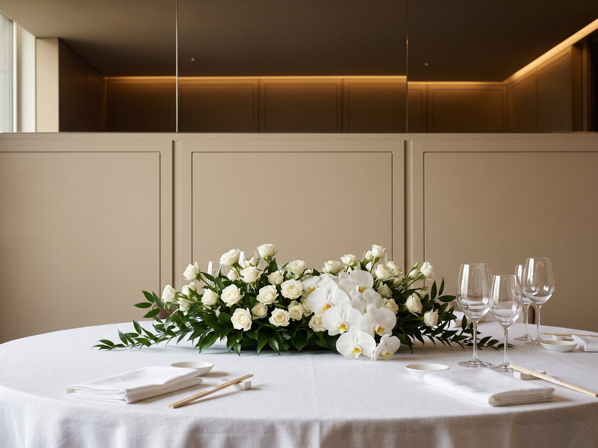 Arranjo baixo e alongado de rosas brancas, orquídeas phalaenopsis brancas e folhagem verde-escura no centro de uma mesa redonda posta com toalha de linho branco, taças de cristal, guardanapos dobrados e pratos de porcelana, numa sala privativa de restaurante: atrás, o lambril de painéis bege e a faixa de espelho que reflete o forro escuro e a luz quente embutida.