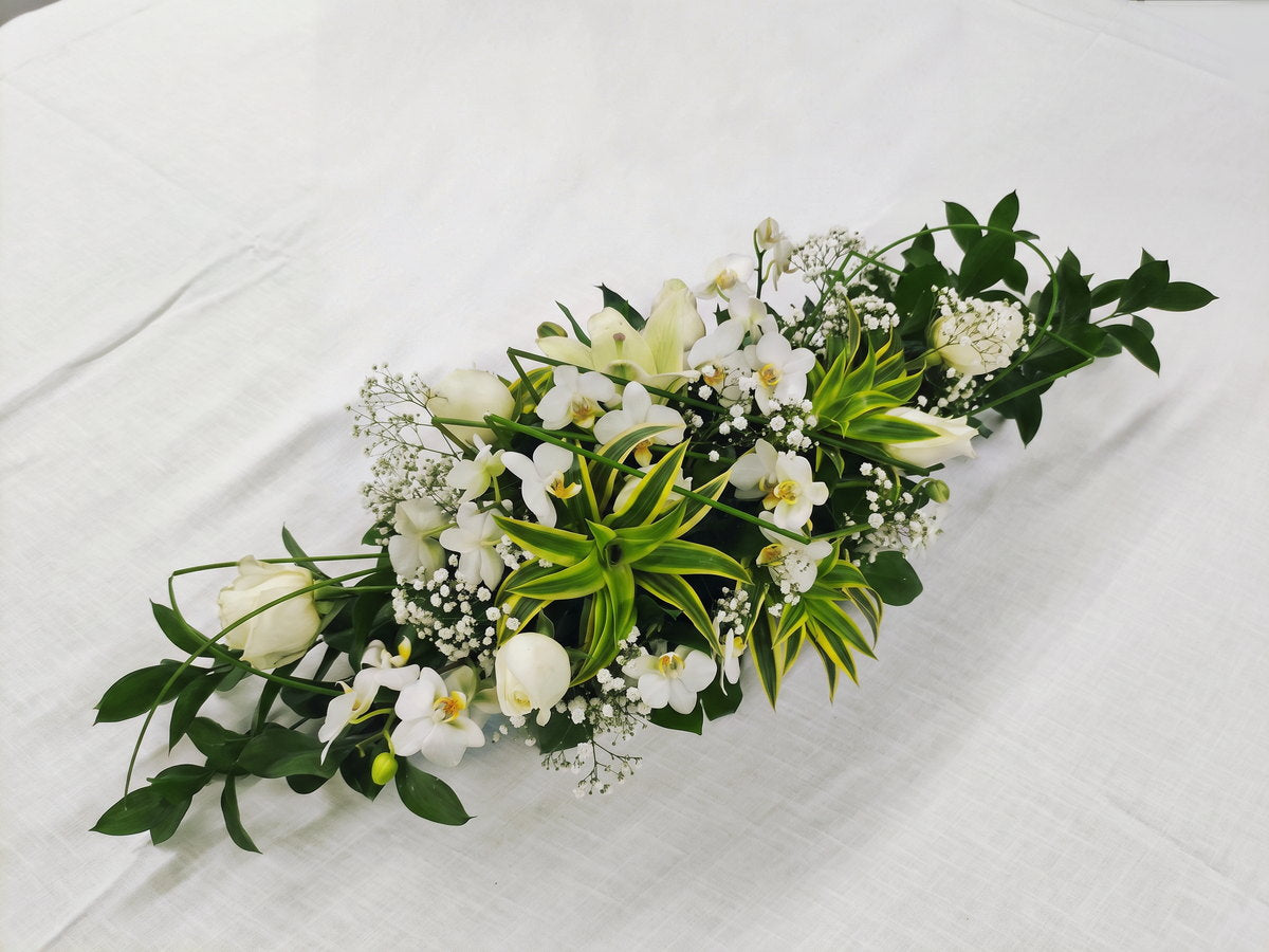 Vista de cima de um arranjo baixo e alongado no centro de uma mesa redonda com toalha de linho branco: rosetas de dracena de folha verde com borda creme, orquídeas phalaenopsis brancas, lírios, rosas brancas e nuvens de mosquitinho, com lâminas finas de capim cruzando por cima.