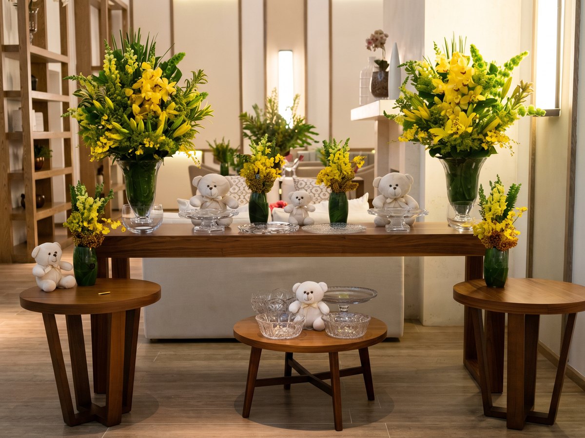 Mesa de evento montada pela casa num lounge claro: sobre o aparador de nogueira, dois arranjos altos de orquídeas cymbidium amarelas, lírios, molucela verde e solidago em vasos de vidro com folha por dentro, e entre eles arranjos pequenos de oncídio amarelo em vasinhos verdes, ursinhos de pelúcia brancos e travessas de cristal lapidado. À frente, três mesinhas redondas de nogueira com mais ursinhos, cristais e arranjos pequenos. A luz do dia entra pela direita e deita as sombras das mesas no piso.