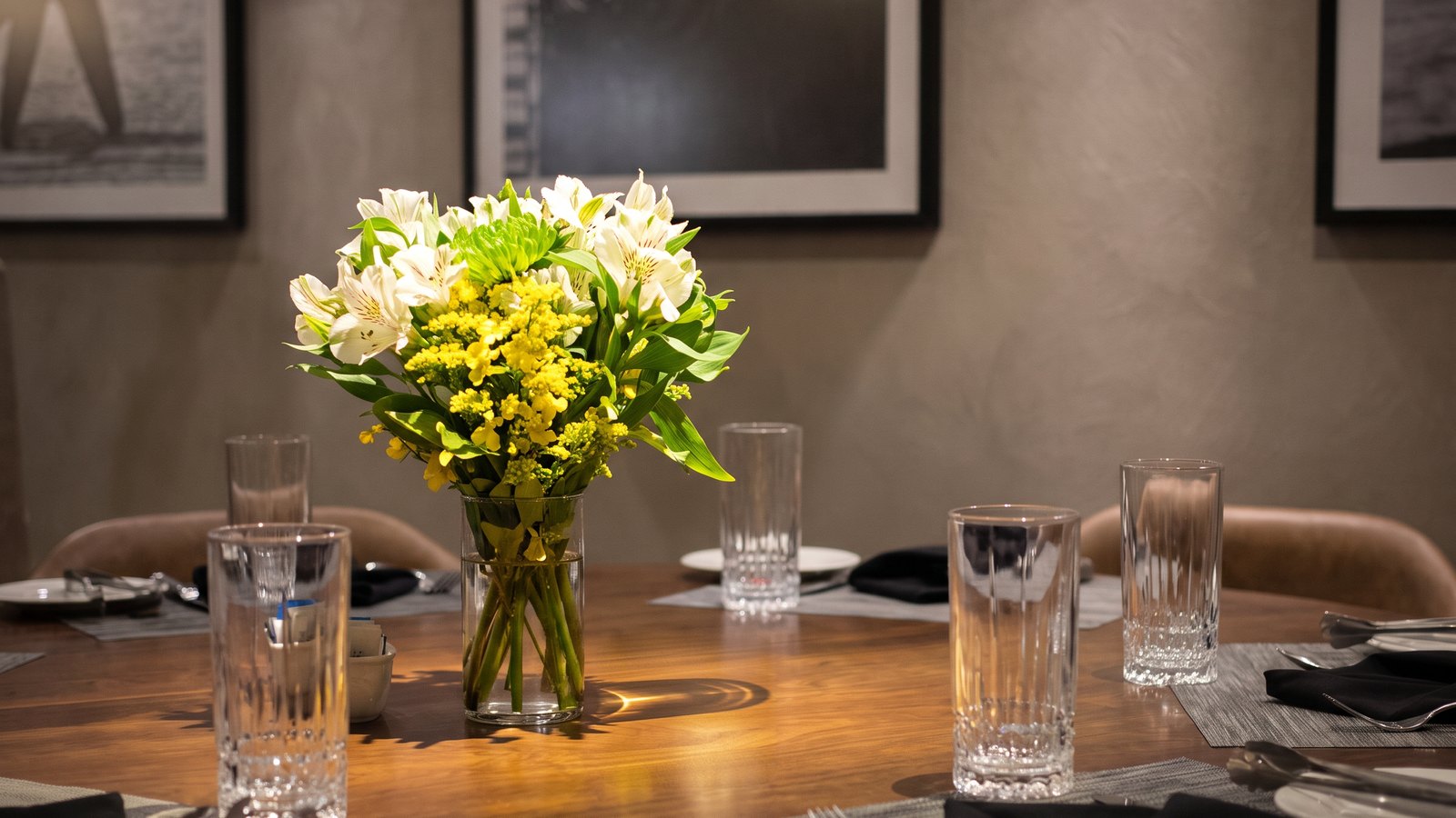 Num jantar corporativo em sala privativa, o arranjo baixo da casa no centro da mesa redonda de madeira: alstroemérias brancas, solidago amarelo e folhas verdes num cilindro de vidro. Um foco de luz quente acende o buquê; em volta, copos de cristal, guardanapos pretos e, ao fundo, as cadeiras de couro caramelo e os quadros em preto e branco na parede, fora de foco.
