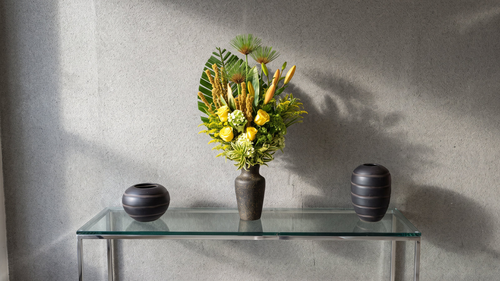 Na recepção de um cliente, o arranjo da casa sobre o aparador de vidro e aço, entre os dois vasos escuros de cerâmica: rosas amarelas, botões de lírio alaranjados ainda fechados, hortênsia verde e branca, solidago e espigas secas cor de palha, com leques de palmeira e cabeças de papiro abrindo por cima, num vaso escuro salpicado de ouro. A luz entra rasante pela esquerda, acende o granito da parede e joga a sombra do arranjo para a direita.